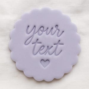 May include: A light purple circular cookie stamp with a scalloped edge. The centre features the embossed text "your text" in a cursive font, positioned above a small, simple heart shape. The design is pressed into a smooth, flat surface.