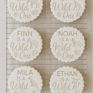 May include: Six round white scalloped biscuits featuring embossed text and animal silhouettes. Each biscuit displays a name followed by the phrase "IS A Wild One" and a tropical leaf design. The biscuits are arranged on a metal cooling rack.