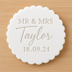 May include: A white, scalloped-edge wedding coaster with the text "MR & MRS Taylor 18.09.24" in a light grey script. The coaster is set against a light brown wooden surface.