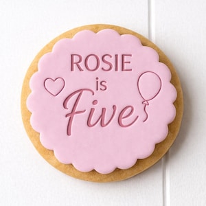 May include: A decorated biscuit with a pink icing layer. The icing is scalloped and features the text "Rosie is Five", a heart, and a balloon. The biscuit has a light brown, baked appearance.
