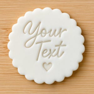May include: A circular white cookie stamp with a scalloped edge featuring the embossed text Your Text in a cursive script font, positioned above a small embossed heart shape centred at the bottom of the circular surface.