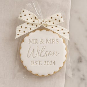 May include: A round scalloped biscuit with white icing, featuring the embossed text MR & MRS Wilson EST. 2024. The biscuit is sealed in a clear plastic bag and tied at the top with a cream-coloured ribbon featuring a gold polka dot pattern.