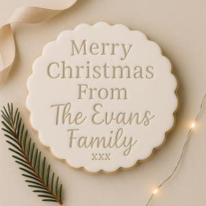 May include: A round, scalloped-edge biscuit with a smooth white icing surface. The text "Merry Christmas From The Evans Family" is embossed in the centre, followed by "xxx" at the bottom. The biscuit is decorated with a pine sprig and string lights.
