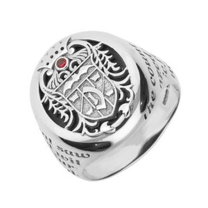 May include: A silver ring with an ornate crest design. The crest features a shield with a letter and is topped with a small red gemstone. The ring has engraved text around the band.