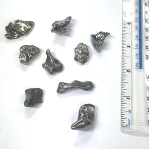 May include: A selection of irregularly shaped, metallic grey meteorites. The meteorites have varying sizes and textures, some with rough, pitted surfaces. A ruler with centimetre measurements is in the background for scale.