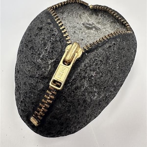 May include: A dark grey stone sculpture with a brass zipper detail. The zipper is partially open, revealing a lighter grey interior. The sculpture has a rough, textured surface and a natural, organic shape. The zipper pull has the text "YKK" on it.