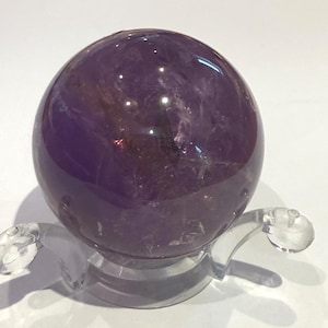 May include: A spherical amethyst crystal ball, displaying a rich purple colour with internal crystalline patterns. The sphere is presented on a clear, curved acrylic stand, giving a floating appearance. Suitable for home decor or crystal collectors.