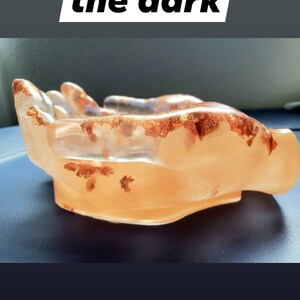 May include: A clear resin hand-shaped bowl with copper flakes embedded in the resin. The bowl is on a blue surface. The text "They glow in the dark" is visible above the bowl.