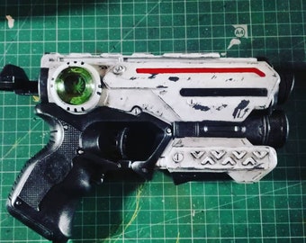 May include: A detailed toy gun in white and black, with a green lens and a red stripe. The design includes intricate patterns and a futuristic aesthetic, possibly for cosplay or themed play. The gun's style suggests a sci-fi theme.