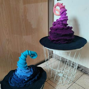 Könnte beinhalten: Zwei gehäkelte Hexenhüte. Einer ist lila mit einer rosa und orangefarbenen Spirale, der andere ist blau und türkis. Beide haben schwarze Krempen. Die Hüte sind handgefertigt und stehen auf einem weißen Drahttisch und einem Metalltablett.