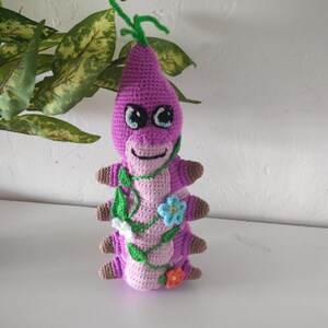 May include: A crocheted purple caterpillar toy with a green stem on top. The caterpillar has brown legs, a smiling face, and is decorated with pink vines, green leaves, and colourful flowers.