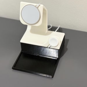 May include: White and black 3D printed charging stand for an Apple Watch. The stand has two charging spots, one for the watch and one for the charging cable.