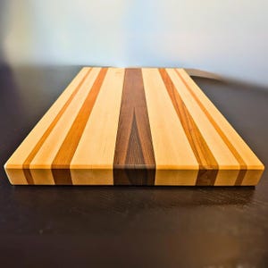 May include: A rectangular wooden chopping board with a variety of wood tones. The board features alternating stripes of light, medium, and dark brown wood. The chopping board is approximately 46 cm long and 30 cm wide.