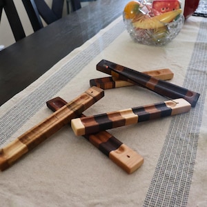 Può includere: Diversi porta incenso in legno in varie tonalità di marrone e beige sono disposti su un runner da tavolo color crema. I supporti rettangolari hanno un'incisione per i bastoncini di incenso. Alcuni hanno un motivo a scacchiera.