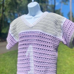 Puede incluir: Top de crochet hecho a mano en tonos lavanda, blanco y verde salvia. El top con cuello en V presenta un dise&ntilde;o a rayas con detalles calados. La prenda de manga corta se exhibe en un maniqu&iacute; blanco.
