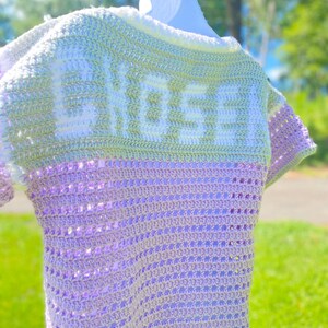 Puede incluir: Un top de ganchillo hecho a mano en tonos lavanda, verde y blanco, colgado de una percha blanca. La palabra "CHOSEN" est&aacute; tejida en la espalda en blanco. El top presenta un dise&ntilde;o de manga corta y un patr&oacute;n de puntada texturizada.