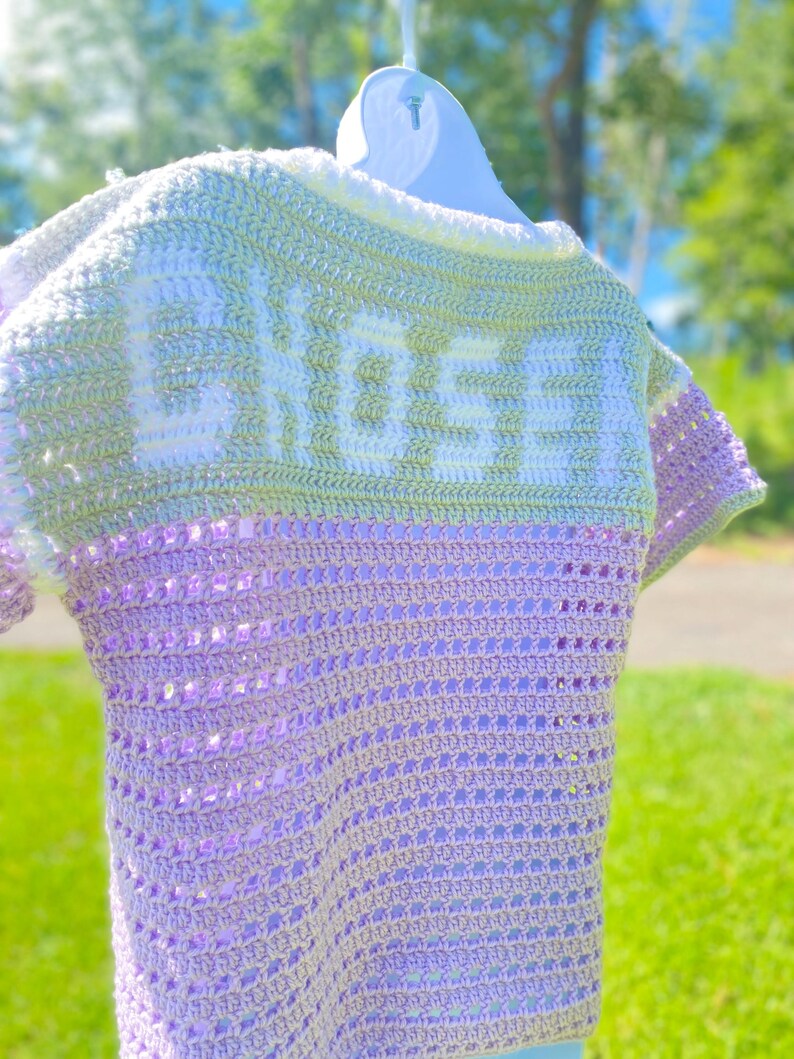 Puede incluir: Un top de ganchillo hecho a mano en tonos lavanda, verde y blanco, colgado de una percha blanca. La palabra "CHOSEN" est&aacute; tejida en la espalda en blanco. El top presenta un dise&ntilde;o de manga corta y un patr&oacute;n de puntada texturizada.