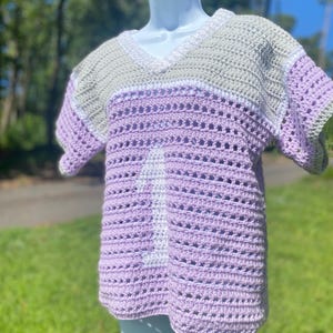 Puede incluir: Una camisa de crochet hecha a mano en tonos lavanda, gris y blanco. La parte superior con cuello en V presenta un dise&ntilde;o a rayas con el n&uacute;mero 1 blanco en la parte delantera. La camisa se exhibe en un maniqu&iacute; blanco al aire libre.
