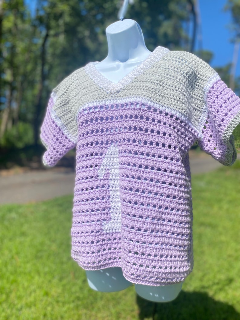 Puede incluir: Una camisa de crochet hecha a mano en tonos lavanda, gris y blanco. La parte superior con cuello en V presenta un dise&ntilde;o a rayas con el n&uacute;mero 1 blanco en la parte delantera. La camisa se exhibe en un maniqu&iacute; blanco al aire libre.