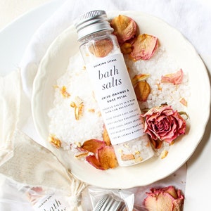 May include: Two small glass bottles filled with bath salts and dried rose petals. The bottles have white labels with the text "bath salts" and "sweet orange & rose petal". The bottles are sitting on a white plate with dried rose petals. A white card with black calligraphy says "Take one! From our shop to yours ♡ xo".