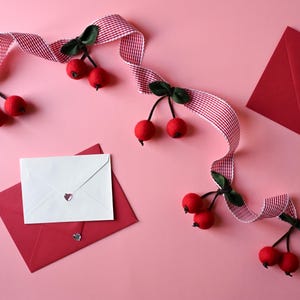 May include: A pink background with red envelopes, a white envelope, and a red and white ribbon adorned with red cherry decorations. The envelopes have heart-shaped embellishments.