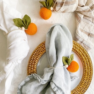 May include: Two white napkins with orange felt fruit napkin rings. One napkin is on a gold charger with a blue napkin. The orange felt fruit napkin rings have green felt leaves.