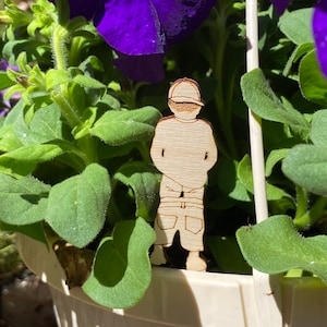 Może przedstawiać: Drewniana wycinanka przedstawiająca osobę w kapeluszu i spodniach, stojącą w doniczce z fioletowymi petuniami.