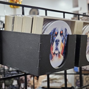 以下が含まれることがあります： 水彩画で描かれた犬のイラストが施された丸いコースター4枚セット。コースターは黒い金属製のラックに飾られています。犬はベルニーズ・マウンテンドッグ、ゴールデンレトリバー、ブルドッグ、ボクサーです。