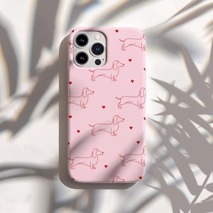 Könnte beinhalten: Rosafarbene Handyhülle mit einem sich wiederholenden Muster aus roten Dackel-Illustrationen und kleinen roten Herzen. Die Hülle hat eine glatte Oberfläche und einen Kameraausschnitt. Das Design ist schlicht und verspielt.