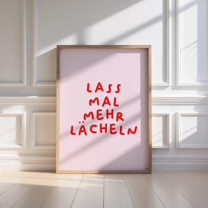 Op de afbeelding: Een ingelijste poster met de Duitse zin "Lass mal mehr lächeln" in rode, handgeschreven letters op een roze achtergrond. De lijst is lichtbruin en de poster leunt tegen een witte muur.