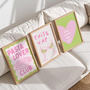 May include: Three framed art prints with playful designs. One says "Pasta Lover Club" with a pasta illustration, another says "This & Yap" with wine glasses, and the last says "How Lucky Are We?" with a heart. Each print has a light green border and a wooden frame.