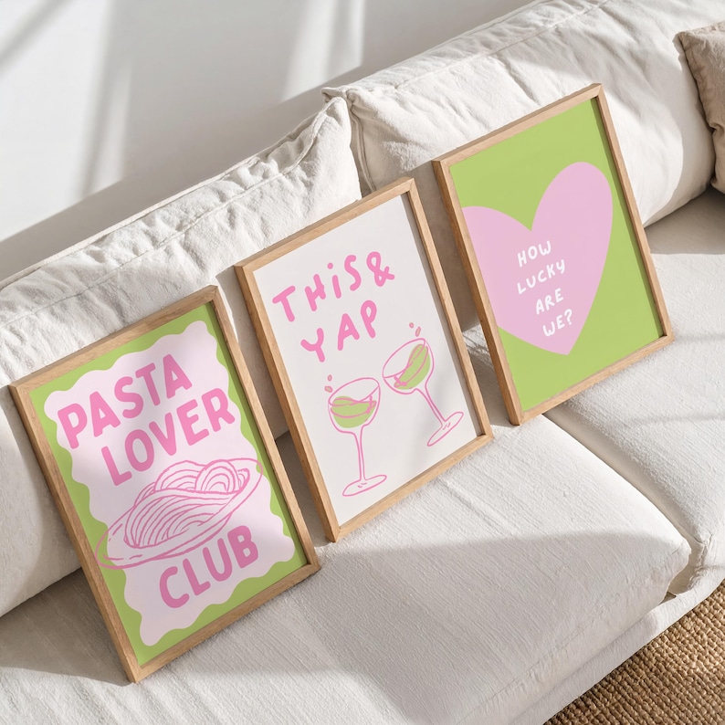 May include: Three framed art prints with playful designs. One says "Pasta Lover Club" with a pasta illustration, another says "This & Yap" with wine glasses, and the last says "How Lucky Are We?" with a heart. Each print has a light green border and a wooden frame.