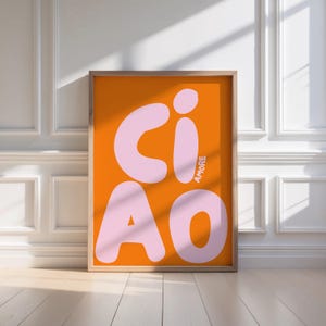 Op de afbeelding: Ingelijste kunstdruk met het woord "CIAO" in grote, lichtroze letters op een oranje achtergrond. Het woord "AMORE" is verticaal in de rechter benedenhoek gedrukt. De lijst is lichtbruin.