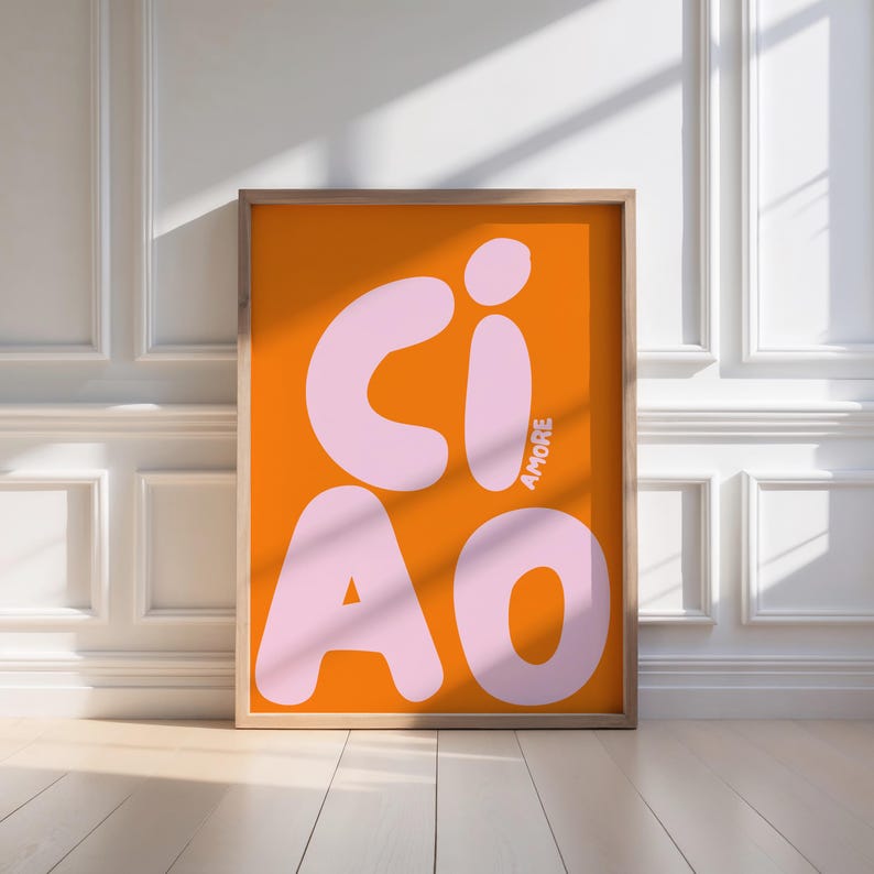 Op de afbeelding: Ingelijste kunstdruk met het woord "CIAO" in grote, lichtroze letters op een oranje achtergrond. Het woord "AMORE" is verticaal in de rechter benedenhoek gedrukt. De lijst is lichtbruin.