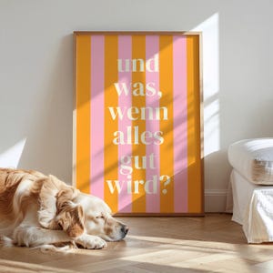 Könnte beinhalten: Ein gerahmtes Poster mit vertikalen orangefarbenen und rosa Streifen, das den deutschen Text "und was, wenn alles gut wird?" in Weiß zeigt. Ein Golden Retriever liegt vor dem Poster auf dem Boden.