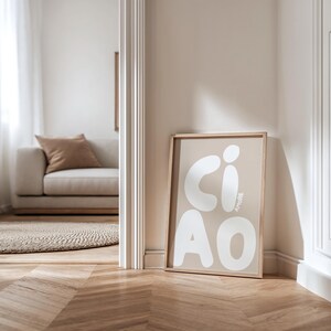 K&ouml;nnte beinhalten: Ein gerahmter Kunstdruck mit dem Wort "CIAO" in gro&szlig;en, wei&szlig;en, abgerundeten Buchstaben auf beigem Hintergrund. Das Wort "AMORE" ist vertikal in kleineren wei&szlig;en Buchstaben geschrieben. Der Druck lehnt an einer Wand in einem Raum mit Sofa und Teppich.