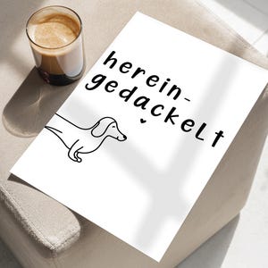 May include: A white print featuring a black line drawing of a dachshund and the German phrase "herein-gedackelt". A glass of coffee sits to the left of the print. The print is on a beige surface.