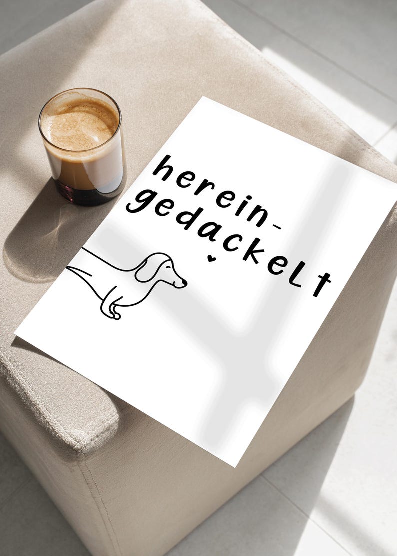May include: A white print featuring a black line drawing of a dachshund and the German phrase "herein-gedackelt". A glass of coffee sits to the left of the print. The print is on a beige surface.