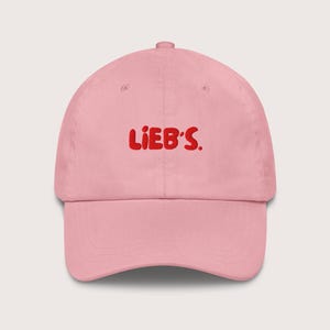 Pode incluir: Boné de beisebol rosa claro com a palavra "LIEB'S" em letras vermelhas. O boné tem uma aba curva e uma alça ajustável na parte de trás. O chapéu é feito de um tecido macio e durável.