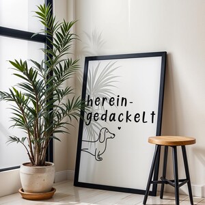May include: A framed art print with the words "herein-gedackelt" and a dachshund illustration. The black frame surrounds a white background. A potted plant and a wooden stool with black legs are also in the image.