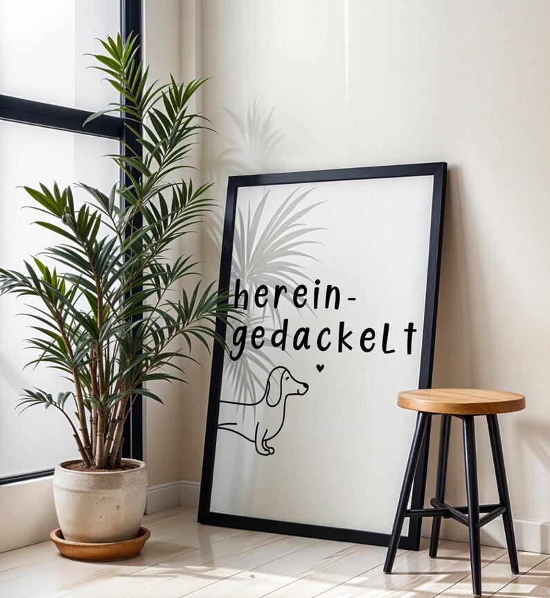 May include: A framed art print with the words "herein-gedackelt" and a dachshund illustration. The black frame surrounds a white background. A potted plant and a wooden stool with black legs are also in the image.