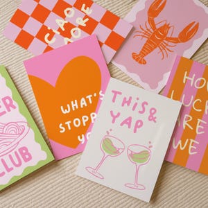 May include: A collection of colorful greeting cards with various designs and text. The cards feature a pink and orange color scheme, with illustrations of a lobster, wine glasses, and a heart. Some cards have text like "Ciao Amore" and "This & Yap."