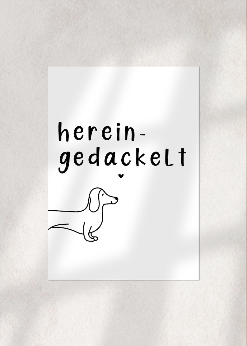 May include: A white print featuring a black line drawing of a dachshund dog and the German phrase "herein-gedackelt" in black text. A small black heart is above the dog. The image is on a white background.