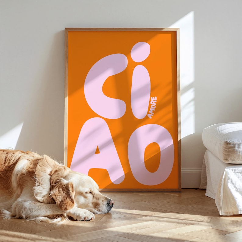 Op de afbeelding: Een ingelijste oranje kunstprint met het woord "CIAO" in grote, lichtroze letters. Het woord "AMORE" staat verticaal aan de rechterkant. Een golden retriever ligt op de vloer voor de print.
