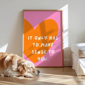 Könnte beinhalten: Ein gerahmter Kunstdruck mit rosa Hintergrund und einer orangefarbenen Herzform. Der Text "IT ONLY HAS TO MAKE SENSE TO YOU" ist in Weiß. Ein Golden Retriever liegt vor dem Kunstwerk auf dem Boden.