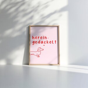 May include: A pink framed print with the text "herein-gedackelt" in red. A red line drawing of a dachshund is below the text.