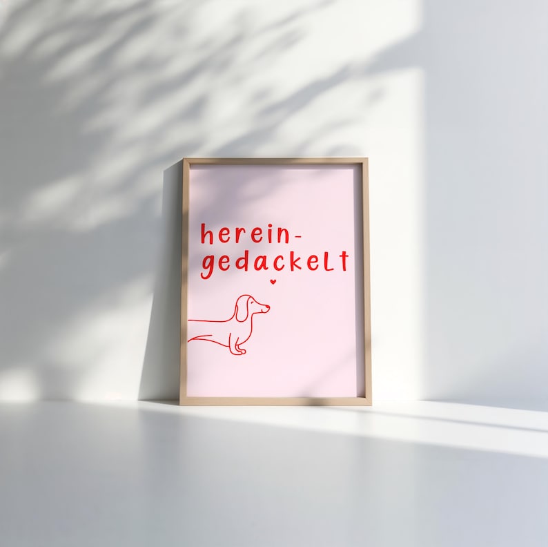 May include: A pink framed print with the text "herein-gedackelt" in red. A red line drawing of a dachshund is below the text.