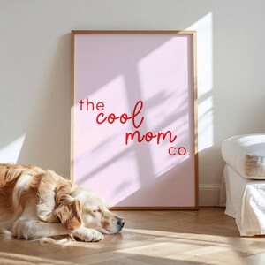 May include: A framed pink print with the text "the cool mom co." in red script. The frame is a light wood colour. A golden retriever dog is lying on the wooden floor in front of the print.