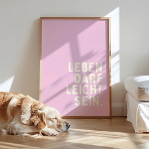 Op de afbeelding: Een roze poster met de Duitse tekst "LEBEN DARF LEICHT SEIN" in een lichte kleur. Een golden retriever ligt op de houten vloer voor de poster.