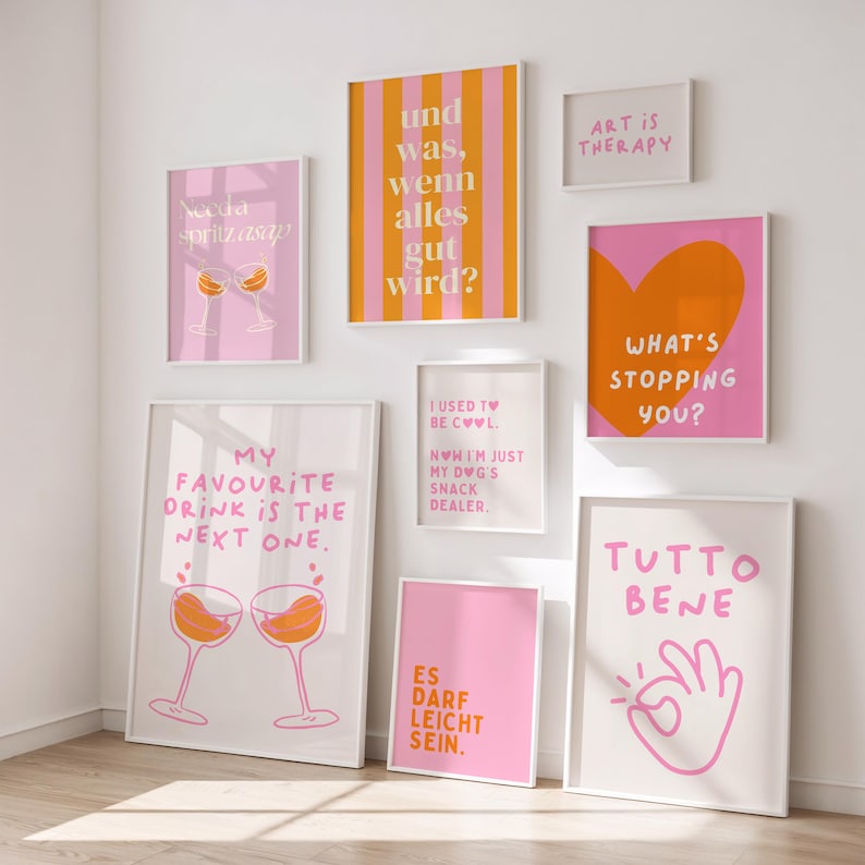 May include: A collection of framed art prints with various designs and text. The prints feature phrases like "What's stopping you?" and "My favourite drink is the next one." Colors include pink, orange, and white. The art is displayed on a white wall.